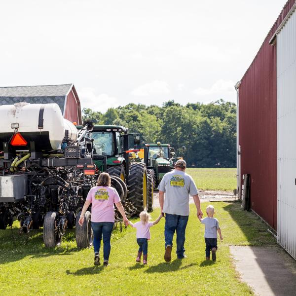 Wide rear shot of Matt and Alisha Gibson and their two children walking between a tractor and a red barn.
