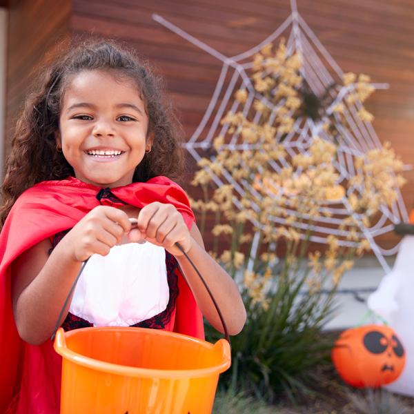 A young girl smiling at the camera while holding a bucket and wearing a cape in front of Halloween decorations.